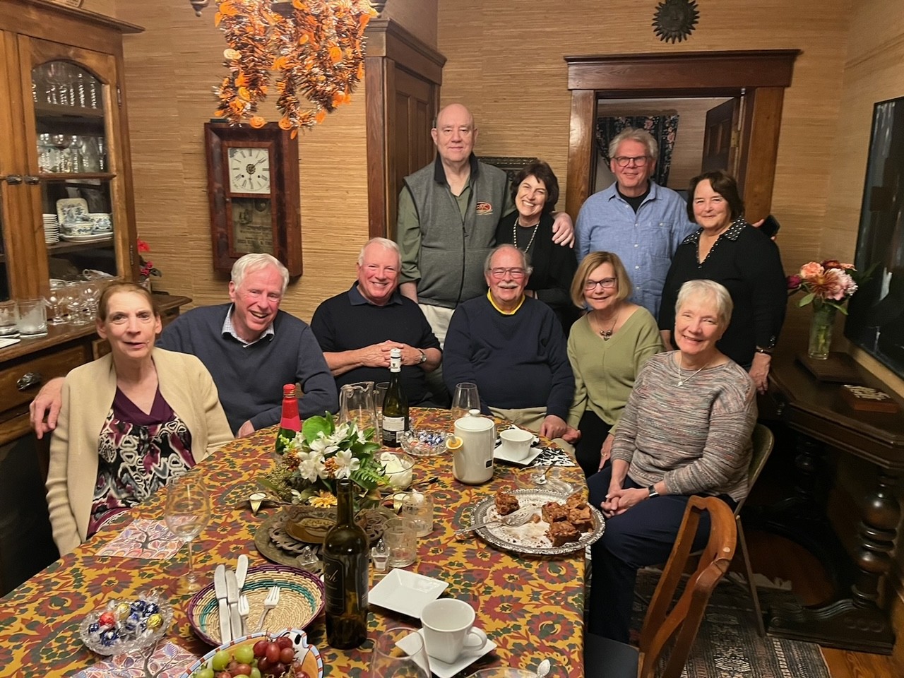 A group of ten smiling individuals gathers around a decorated dining table at a Friday night gathering in Ann Arbor before their 55th reunion at Kalamazoo College in 2024. The front row, from left, includes Elaine Washington, George Washington ’69, Bob Belair ’69, Chuck Meyer ’69, Sandy Bedard Meyer ’67, and Chris Rettich Wilcox ’69, seated with wine glasses, a floral centerpiece, and a spread of food and desserts on a colorful tablecloth. The back row, from left, features Tim Lavalli ’69, Pat Bauer ’69, Tom Matzell ’69, and Holly Belair, standing behind the table. The warmly lit room has wood-paneled walls, a grandfather clock, a china cabinet, and autumn-themed decorations hanging above, with a vase of flowers on a side table.
