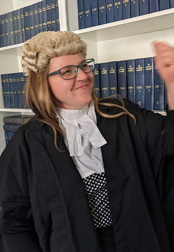 A professional headshot portrait of Claire Madill, a woman with shoulder-length brown hair, smiling gently. She wears a dark blazer over a light pink blouse against a plain white background.