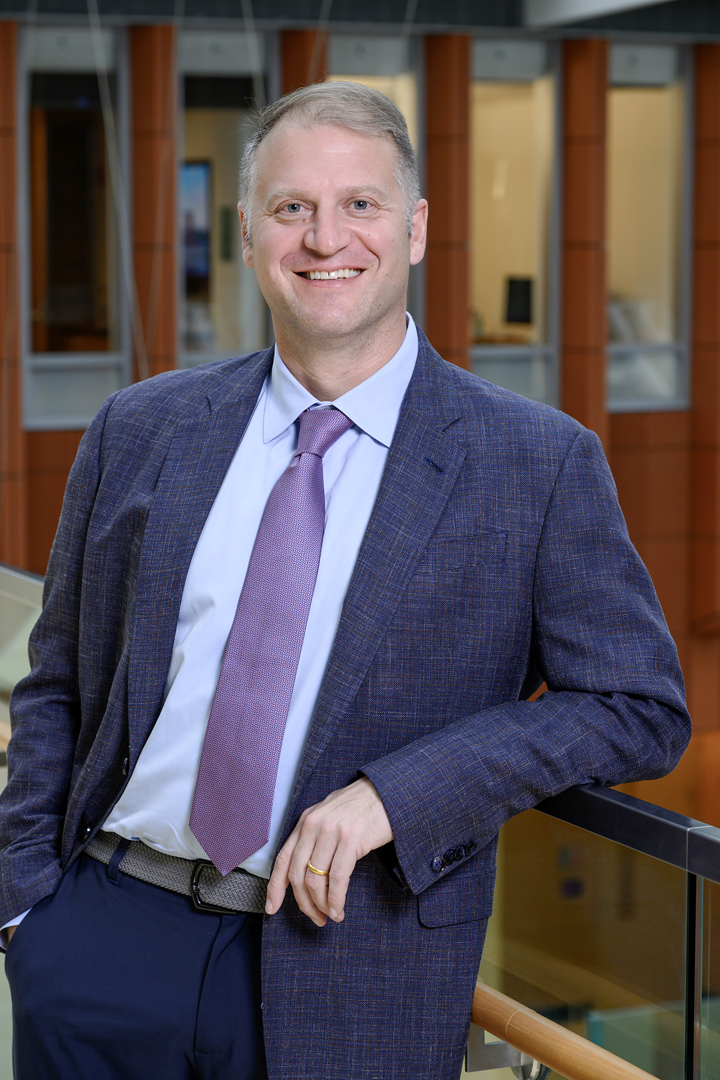 A professional portrait of Ed Mamou ’00, a man with short, light-colored hair, smiling as he leans against a glass and wood railing. He is wearing a blue patterned blazer over a light blue collared shirt and a purple textured tie, paired with dark trousers and a grey woven belt. The background features a modern indoor architectural space with warm wood paneling and large windows.