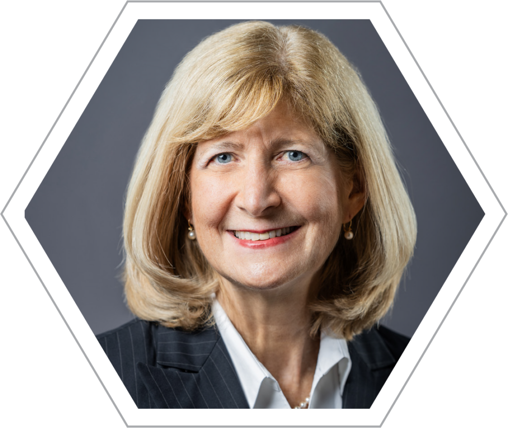 A portrait of Chris Bodurow, smiling directly at the camera. She is wearing a black sleeveless dress and has short, light blonde hair. The setting is indoors, with a warm, slightly blurred background featuring wooden shelves and subtle lighting.he photo is framed within a white and grey hexagonal border.