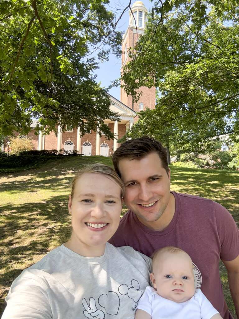A smiling selfie of Joshua Abbott and Allison Tinsey with their infant daughter, Claire Marie, outdoors on a sunny day. Allison is in the foreground wearing a light gray t-shirt, and Joshua is next to her in a maroon t-shirt, holding Claire, who is wearing a white outfit. In the background, Stetson Chapel.