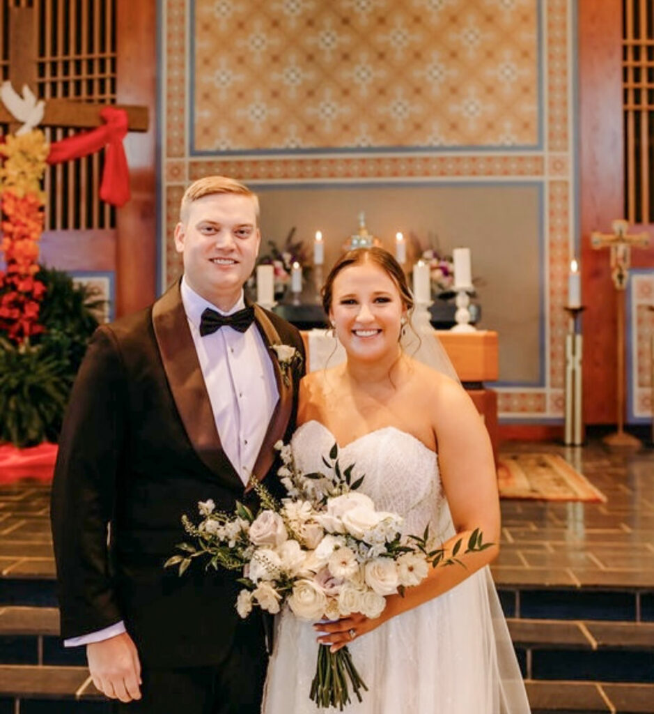A wedding photo of Kaela Van Til and Eric Hart standing together and smiling in a church. Kaela is wearing a white strapless lace wedding dress and holding a bouquet of white and blush roses with greenery. Eric is wearing a black tuxedo with a bow tie. They are positioned in front of an altar decorated with candles and ornate patterned wall hangings.