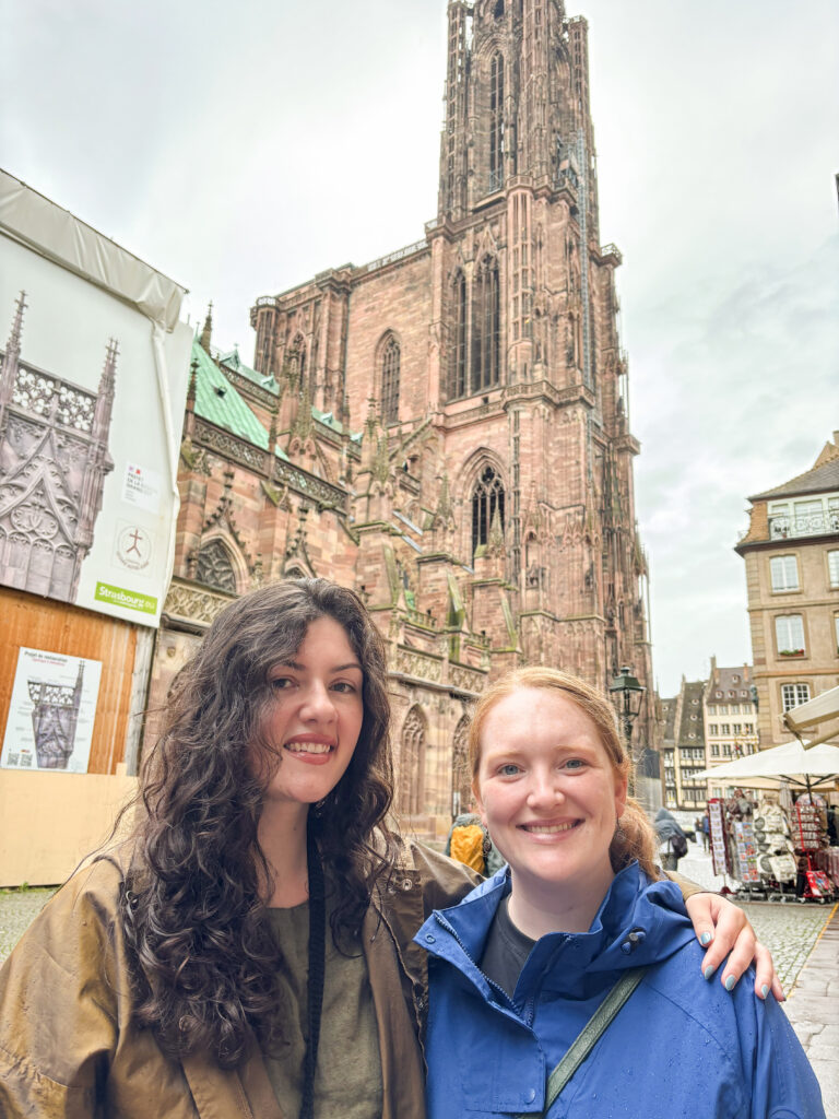A smiling photo of Erica Vanneste-Weber and Shelby Long standing together in Strasbourg, France. Erica, with long dark curly hair and a brown jacket, has her arm around Shelby, who has reddish hair and is wearing a blue rain jacket. They are positioned in a cobblestone square with the towering, intricate Gothic architecture of the Strasbourg Cathedral rising high into the overcast sky behind them. To the left, there is construction scaffolding with a decorative banner, and to the right, a glimpse of European-style buildings and a small outdoor market area.