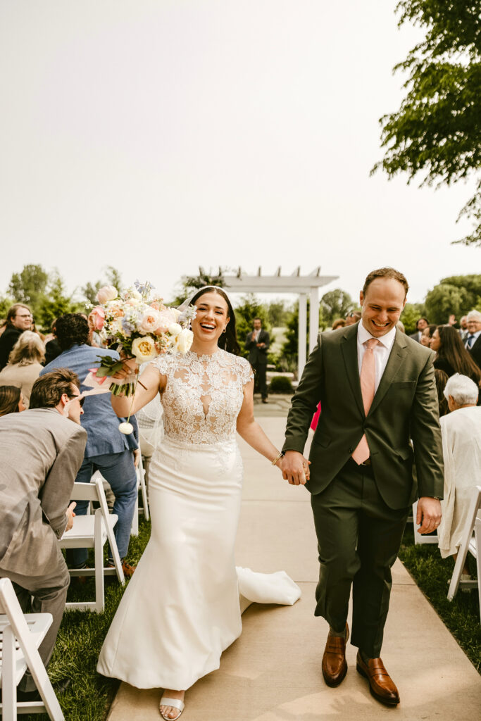A joyful wedding photo of Gabrielle Shimko and David Vanderkloot walking down the aisle together after their ceremony. Gabrielle is laughing radiantly, wearing a white sleeveless gown with an intricate lace bodice and holding up a colorful pastel bouquet. David is smiling beside her, wearing an olive green suit with a light pink tie and brown loafers. They are walking on a paved path lined with guests in white chairs at an outdoor venue, with a white pergola and greenery visible in the background under a bright sky.