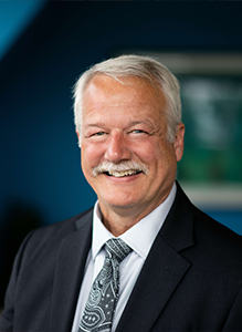 A professional headshot of Keith Crandall ’87, a man with short white hair and a white mustache, smiling warmly at the camera. He is wearing a dark suit jacket over a light blue collared shirt and a grey paisley patterned tie. The background is a vibrant blue wall with a blurred green plant on the left and a soft-focus framed picture on the right.