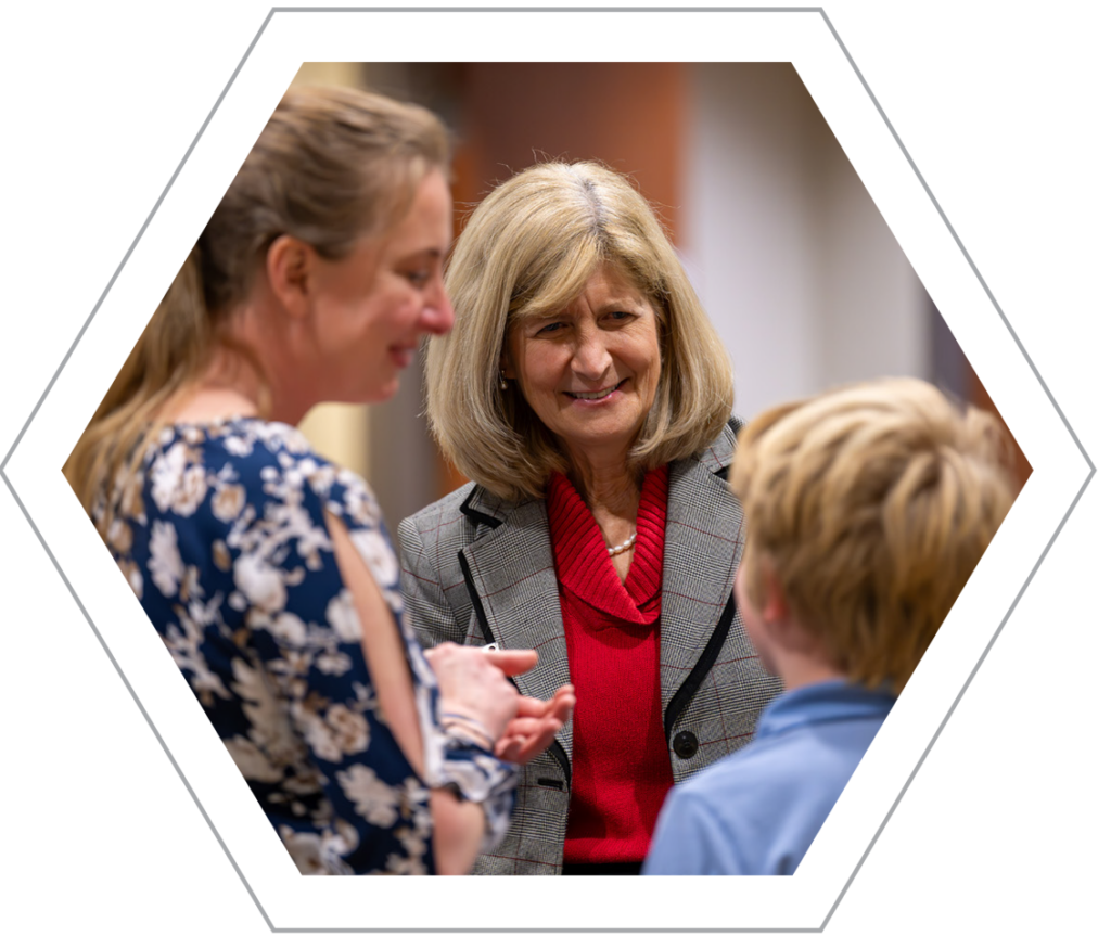 A medium shot of Chris Bodurow in the center, smiling warmly while engaging in conversation with a woman in a floral blouse and a young boy in a blue shirt. Chris Bodurow is wearing a red turtleneck under a grey patterned blazer with black lapel trim and a pearl necklace. The photo is framed within a white and grey hexagonal border.
