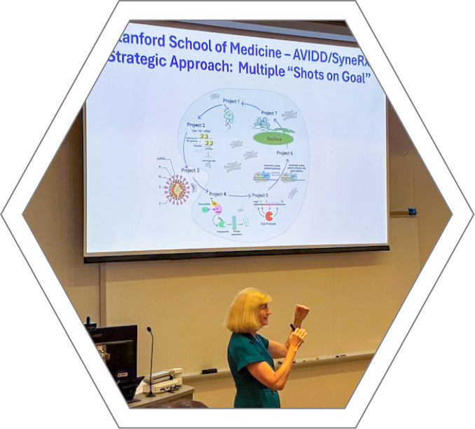 Chris Bodurow gives a lecture at the Indiana University-Indianapolis Department of Chemistry. She stands in the foreground wearing a teal short-sleeved dress, gesturing with her hands while speaking. Behind her, a large projection screen displays a slide titled Stanford School of Medicine – AVIDD/SyneRx Strategic Approach: Multiple "Shots on Goal." The slide features a complex biological diagram showing various research projects mapped around a cellular lifecycle. The image is framed within a white and grey hexagonal border.