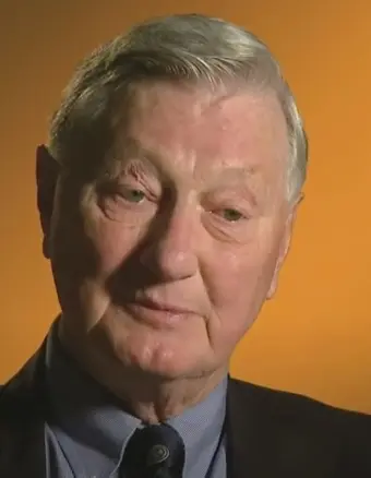 
Gemini said
A close-up portrait of Thomas Cameron Willson from the class of 1952. He is an elderly man with fair skin and short, combed-back grey hair, looking slightly downward with a thoughtful expression. He is wearing a dark suit jacket over a light blue button-down shirt and a dark patterned necktie, set against a solid orange background.