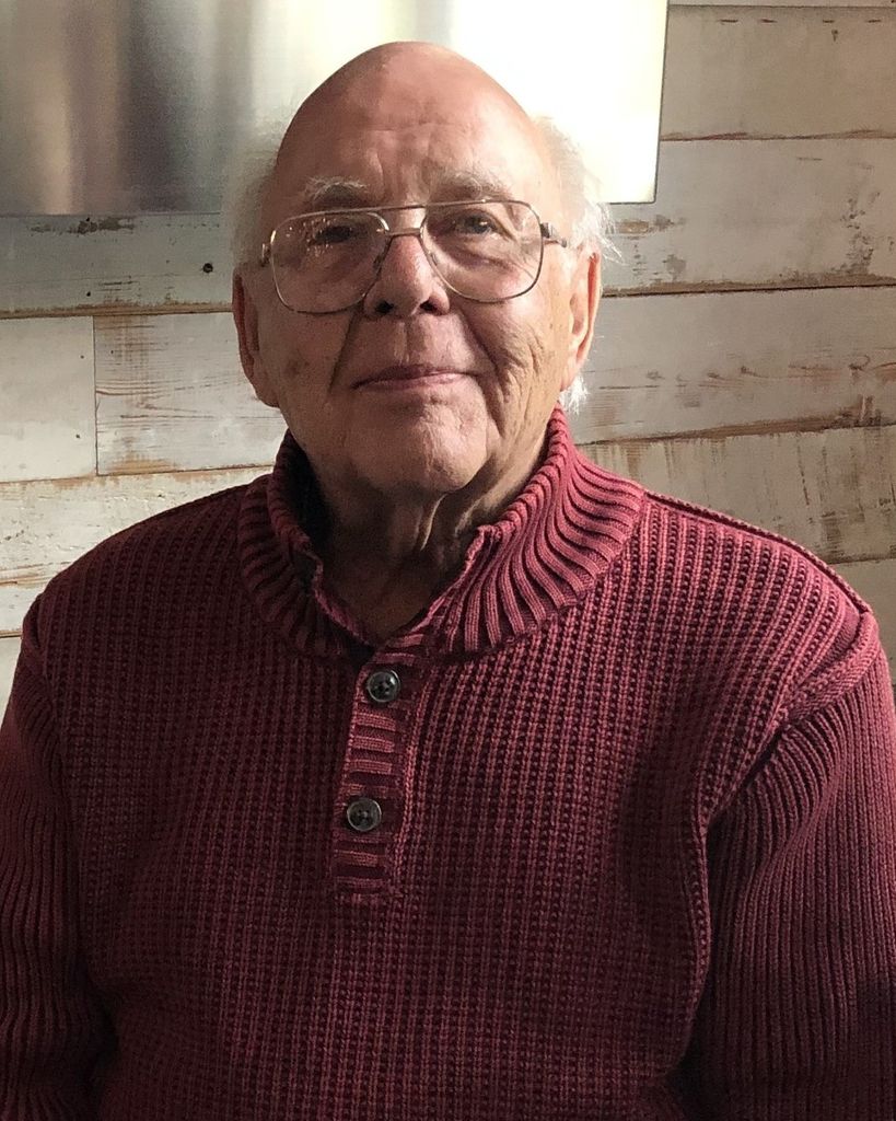 A portrait of Robert Glenn Millard from the class of 1953. He is an elderly man with glasses and a kind expression, shown from the chest up. He is wearing a maroon ribbed henley sweater with two dark buttons, posed against a rustic background of white-washed wooden planks.