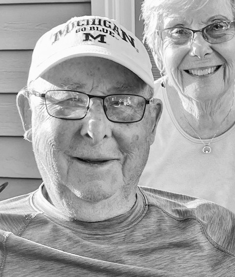 A black-and-white portrait of James "Jim" Harding Fowler from the class of 1956. He is an elderly man with glasses and a friendly smile, wearing a "Michigan Go Blue" baseball cap and a textured athletic shirt. Behind him and slightly to the right, a woman with glasses and a necklace is also smiling for the camera. The background shows the horizontal siding of a house.