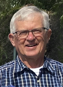 A close-up portrait of Robert “Bob” H. Poel, a man with short white hair and glasses, wearing a blue and white checkered button-down shirt over a white undershirt. He is smiling outdoors with green foliage blurred in the background.
