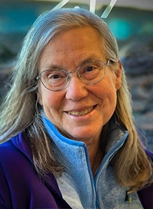 A close-up portrait of Ann Elizabeth Towsley, a woman with long gray hair and glasses, smiling warmly. She is wearing a light blue zip-up fleece jacket over a darker purple garment, with a blurred outdoor or brightly lit background.