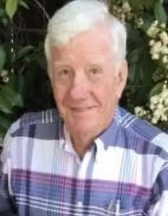 portrait of Mark Robert Millsom from the class of 1959. He is an elderly man with short white hair and a kind, gentle smile. He is wearing a blue, white, and purple plaid button-down shirt, posed outdoors in front of lush green foliage and small white flowers.