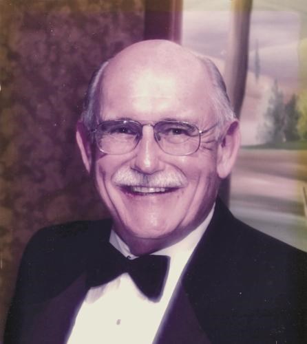 A portrait of Roger Bobertz from the class of 1959. He is an elderly man with glasses and a white mustache, smiling broadly for the camera. He is dressed formally in a black tuxedo with a black bow tie and a white pleated shirt. The background is slightly blurred with warm, indoor lighting.
