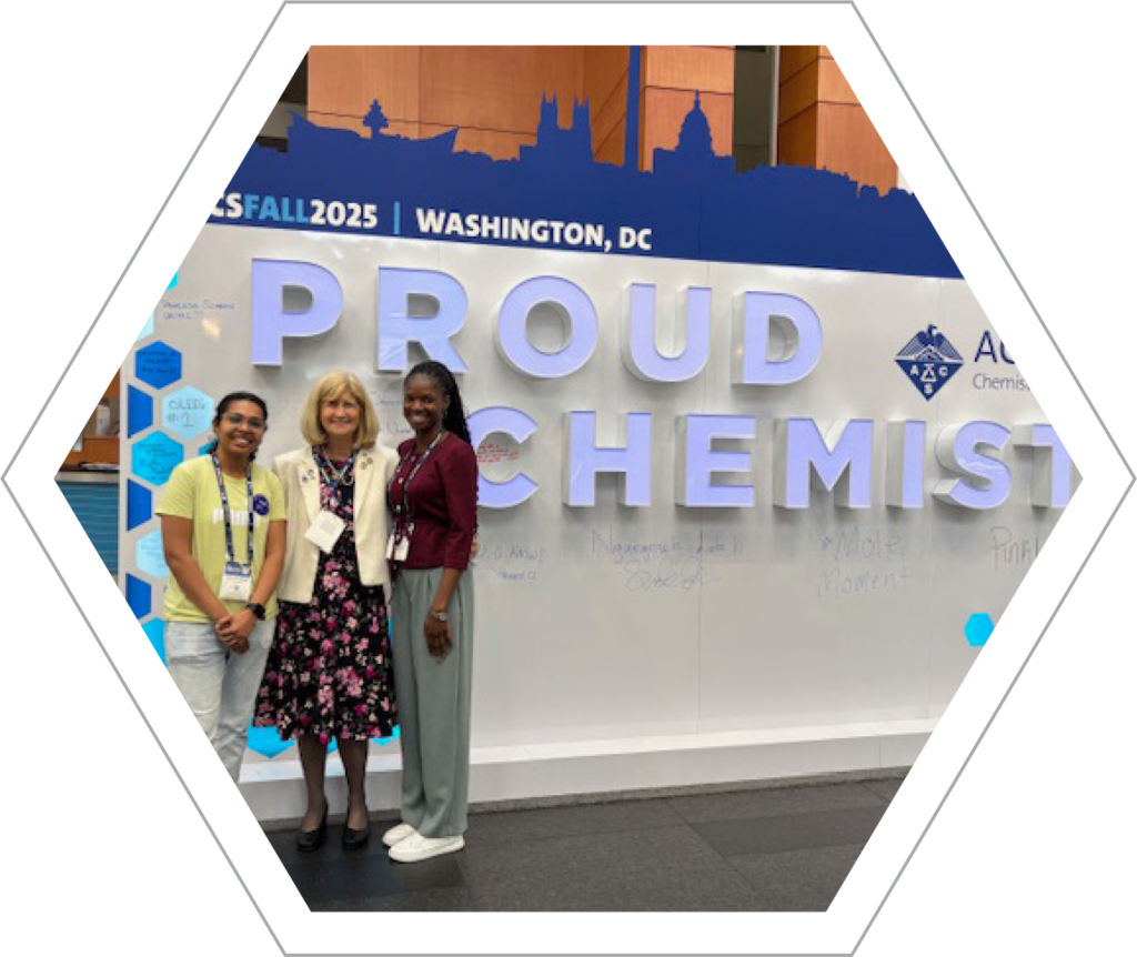  Chris Bodurow stands in the center of two women at the ACS Fall 2025 conference in Washington, D.C. She is wearing a white blazer over a black floral dress. To her left is a woman in a yellow shirt and light-colored jeans, and to her right is a woman in a maroon long-sleeved shirt and grey trousers. They are posing in front of a large white wall featuring the words PROUD CHEMIST in illuminated blue letters, topped with a blue silhouette of the Washington, D.C. skyline. The image is enclosed in a white and grey hexagonal frame.