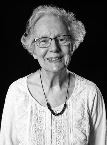 A black-and-white portrait of Barbara (Lape) Zabel from the class of 1961. She is an elderly woman with short, wavy light-colored hair and glasses, smiling warmly at the camera. She is wearing a light-colored lace-detailed blouse and a simple dark beaded necklace, set against a solid black background.