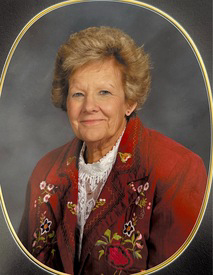 A portrait of Diane (White) Doyle from the class of 1962, framed in a gold oval. She is an elderly woman with short, styled blonde hair and a gentle smile. She is wearing a white lace-trimmed blouse under a distinctive reddish-brown jacket featuring colorful floral embroidery. The background is a neutral, mottled grey.