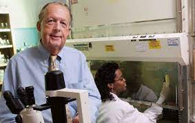 A portrait of Richard “Dick” W. Compans standing in a laboratory setting. He is wearing a light blue dress shirt and a tan tie, positioned next to a microscope. In the background, a colleague in a white lab coat is working inside a laboratory fume hood.