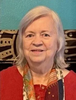 A portrait of LaRay Denzer, a woman with shoulder-length white hair, wearing a red top and a colorful patterned scarf. She is smiling forward against a bright blue wall, with a black and white patterned textile visible behind her shoulders.