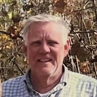 A portrait of Olaf Virro, a man with short white hair, wearing a blue and white checkered button-down shirt. He is smiling slightly toward the camera with a background of autumnal trees and branches.