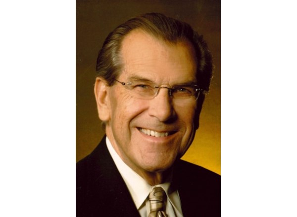 A portrait of Maris Rushevics, a man with swept-back dark hair and glasses, wearing a dark suit jacket, a white dress shirt, and a patterned tie. He is smiling warmly toward the camera against a warm, gradient-lit background.