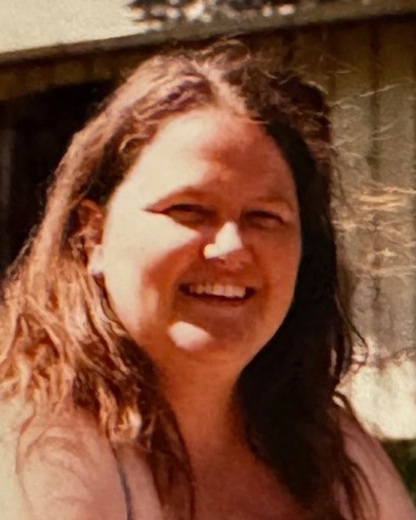 A portrait of Kathryn (Hammer) Kerman, a woman with long, wavy brown hair, smiling brightly toward the camera. The photo is a close-up shot taken outdoors with a soft, natural light and a slightly blurred background.