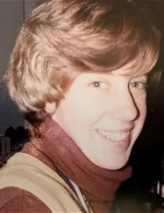 A portrait of Susan (Fake) Habel, a woman with short, wavy reddish-brown hair, smiling in profile. She is wearing a dark reddish-brown turtleneck sweater under a lighter tan garment, with a dark, indistinct background.