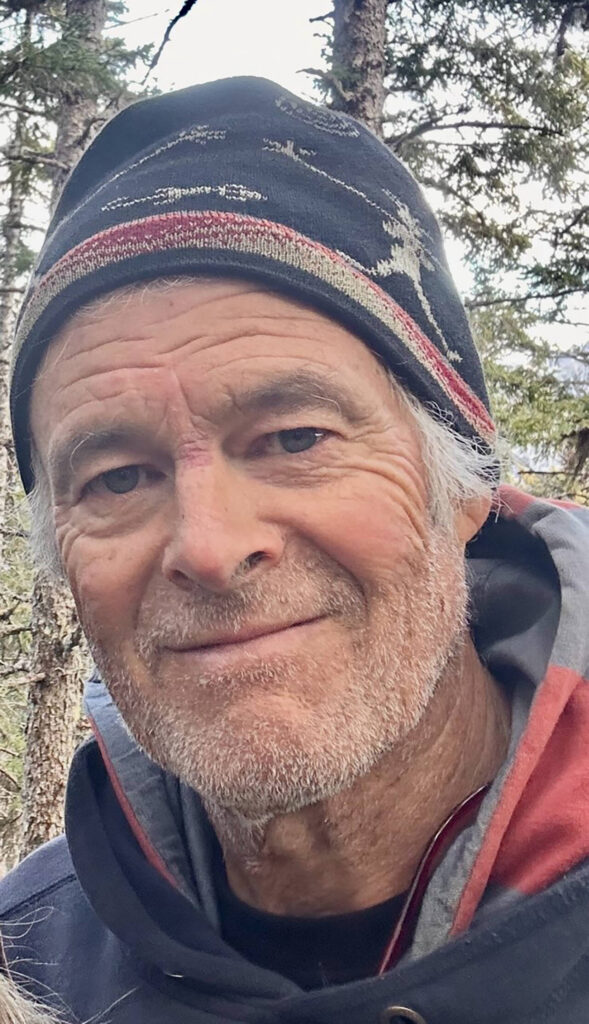 A portrait of David Seaman, a man with a gray beard and short hair, wearing a dark beanie with a red and tan horizontal stripe and a patterned design. He is wearing a dark hoodie and is smiling slightly at the camera with a forest of evergreen trees in the background.