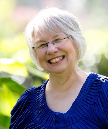 A portrait of Emily Jean Daniel, a woman with short white hair and glasses, smiling broadly at the camera. She is wearing a textured, dark blue short-sleeved top and is positioned outdoors with bright, sunlit green foliage blurred in the background.