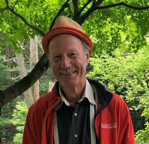 A portrait of Ronald Allen Klug, a man with a mustache and light-colored hair, smiling at the camera. He is wearing a straw fedora with an orange band, a red zip-up jacket over a dark button-down shirt, and light-colored suspenders. He is standing outdoors with lush green trees in the background.