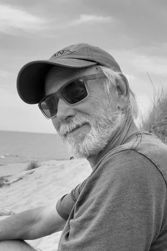 A black and white portrait of Gary Edward Wichman ’74, smiling while sitting on a sandy beach. He is wearing a baseball cap, sunglasses, and a short-sleeved shirt. The background shows beach dunes and the calm surface of a large body of water under a light sky.