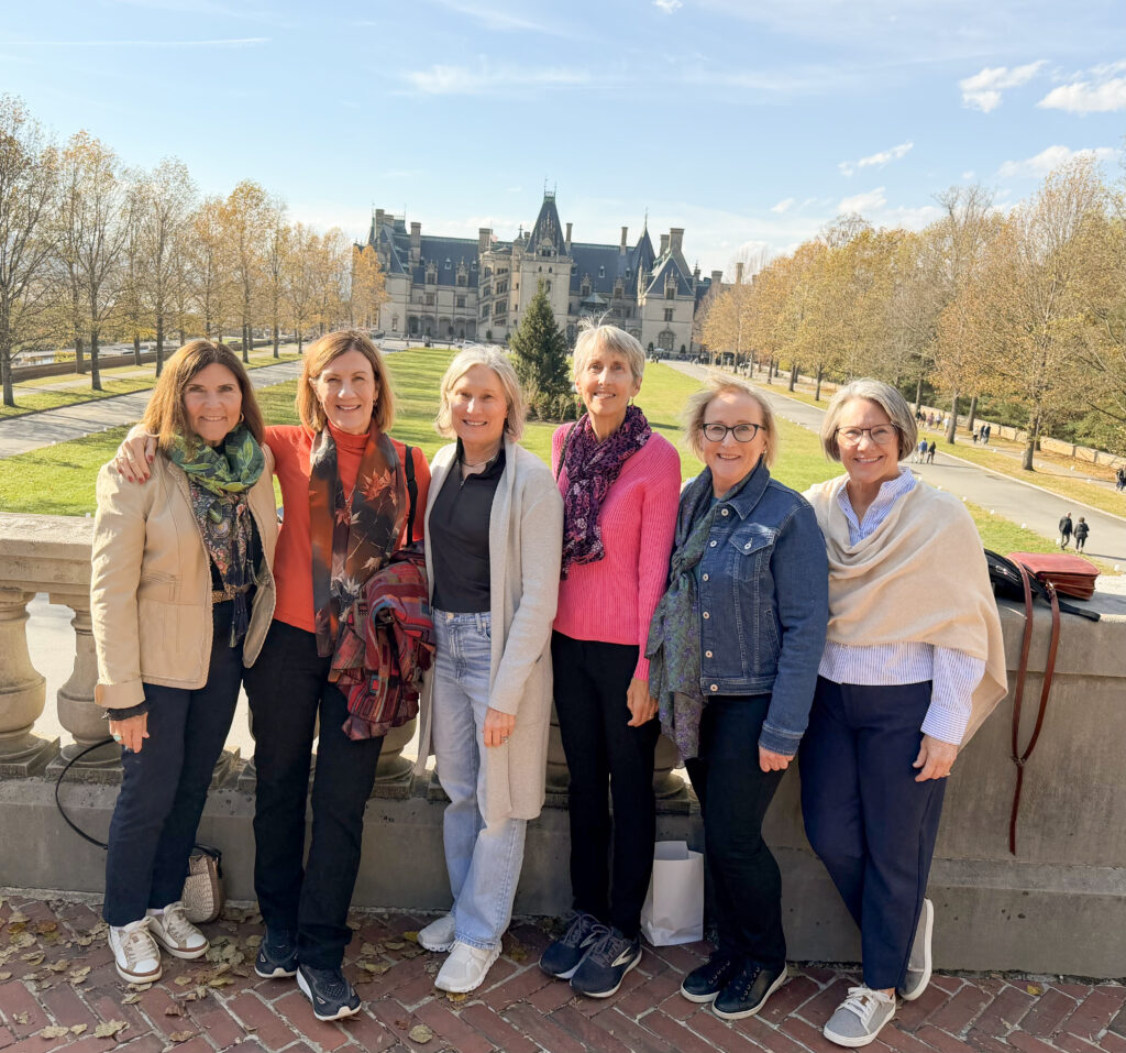 Six women, identified as the Class of 1978 suitemates, stand together for a group photo on a sunny day with a grand, chateau-style estate in the background. They are positioned in a row behind a stone balustrade, smiling warmly. From left to right are Nancy Moody in a tan jacket and green scarf; Karen Gengle in a bright orange top and patterned scarf; Bonnie Forrester in a black top, light grey cardigan, and jeans; Cindy Berry in a vibrant pink sweater and purple scarf; Renee Oshinski in a denim jacket and patterned scarf; and Annette Campbell in a striped blue shirt and cream-colored wrap. The background features a long green lawn leading up to the historic Biltmore Estate under a clear blue sky.
