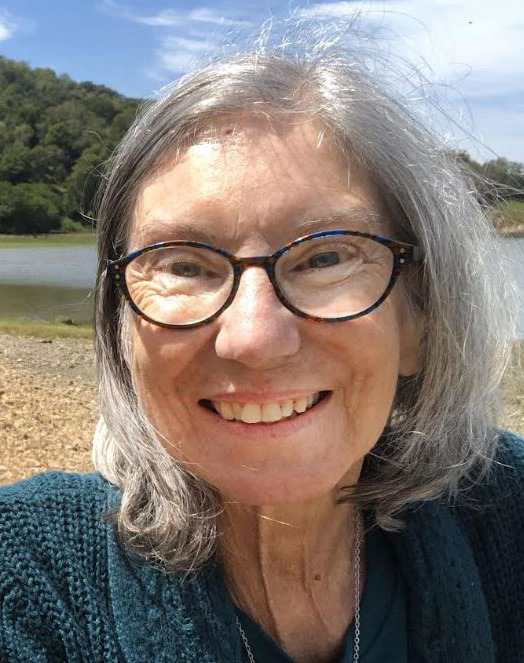A close-up portrait of Eileen E. Varin ’80, a woman with grey hair and glasses, smiling outdoors. She is wearing a dark teal knit sweater and a necklace. The background features a sunny natural setting with a body of water, a shoreline, and a forested hill under a blue sky.