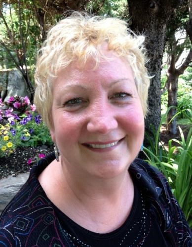 A close-up portrait of Susan (Szabo) Mattano ’81, a woman with short, curly blonde hair, smiling warmly. She is wearing a black top with a patterned jacket or cardigan. The background features a lush garden with green foliage, a tree trunk, and vibrant purple and yellow flowers.
