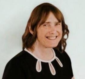 A portrait of Alicia (Cather) Rotgers ’82, a woman with shoulder-length brown hair and bangs, smiling. She is wearing a black top with a decorative white scalloped neckline featuring teardrop-shaped cutouts. The photo is set against a plain, light-colored background.