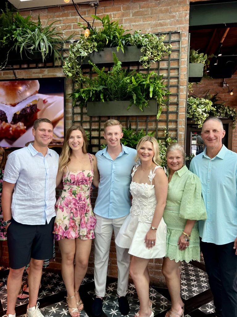 A group of six people posing for a photo in an indoor setting with a rustic-modern aesthetic. Standing from left to right are three young adults followed by an older couple. The group is smiling warmly in front of a brick wall decorated with a vertical garden of lush green ferns and trailing plants. To the left, a digital screen displays a close-up image of a burger. The women are dressed in stylish outfits, including a pink floral dress, a white dress with 3D floral appliqués, and a light green eyelet dress. The men are wearing button-down shirts in shades of light blue and white. The floor features a striking black and white geometric tile pattern.