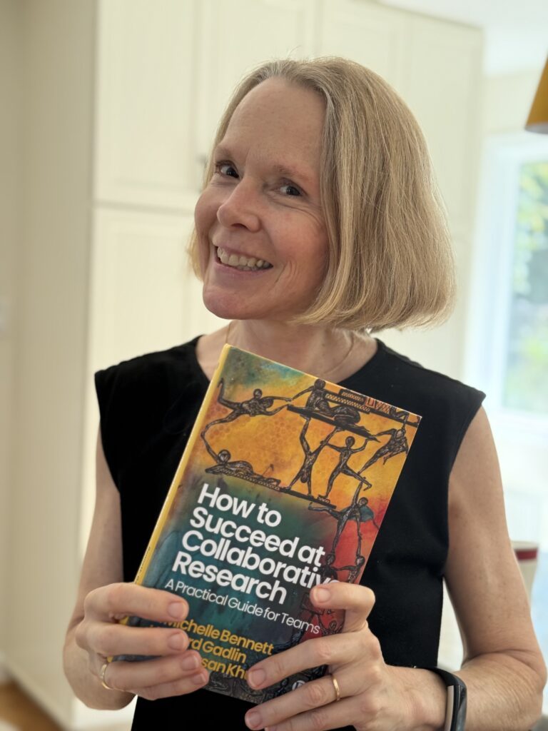 A portrait of L. Michelle Bennett ’85, a woman with short, blonde hair, smiling at the camera and wearing a black, sleeveless top. She is holding a book in both hands. The book is titled "How to Succeed at Collaborative Research: A Practical Guide for Teams" by Michelle Bennett, Gadlin, and San Khou. The book cover features an artistic illustration of figures working together. In the background, there is a blurry kitchen.