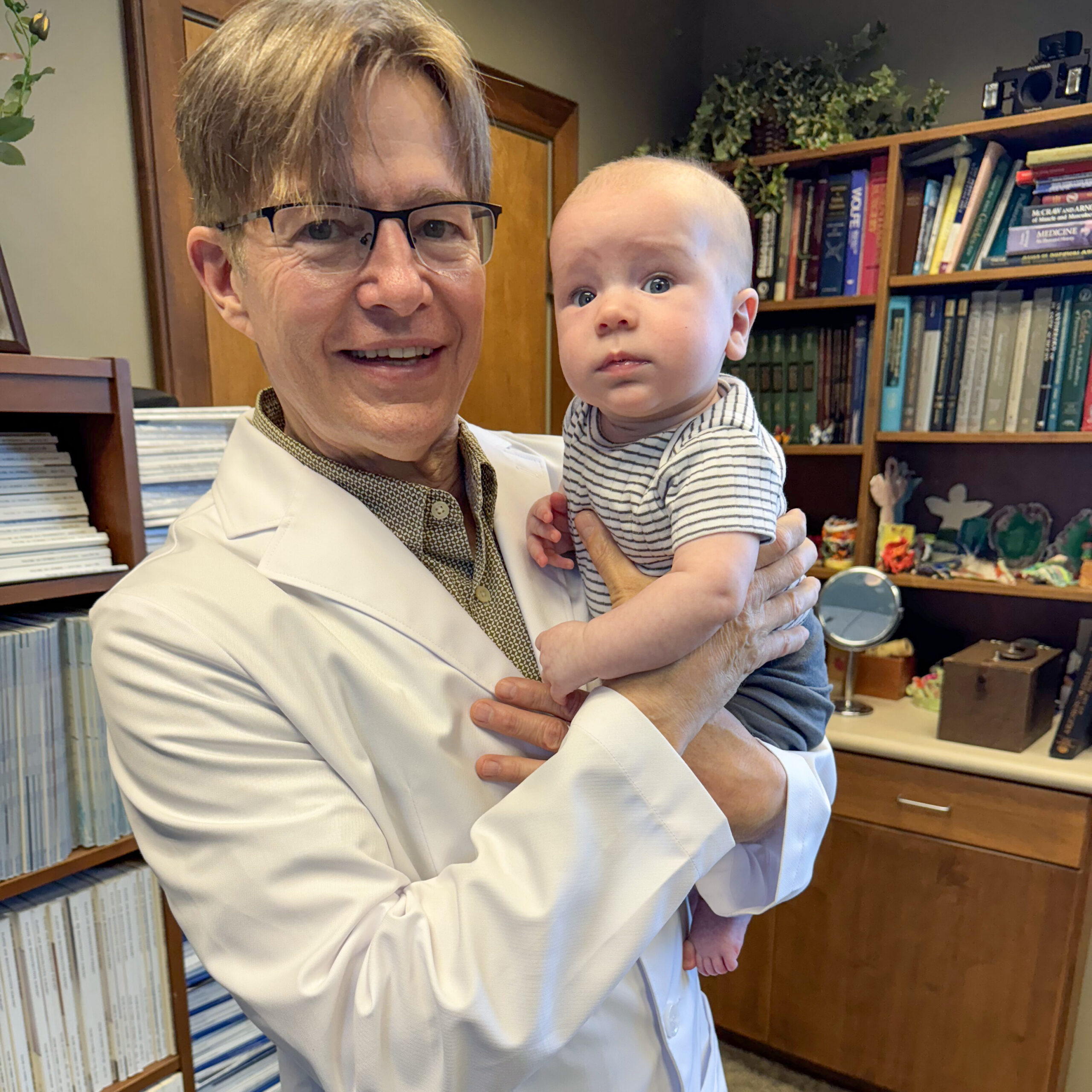 A portrait of Robert Severinac ’85, wearing a white medical lab coat over a patterned collared shirt and glasses, smiling as he holds a baby. The baby is wearing a grey and white striped onesie and looking toward the camera. They are standing in an office or study with a bookshelf filled with medical texts and journals visible in the background.