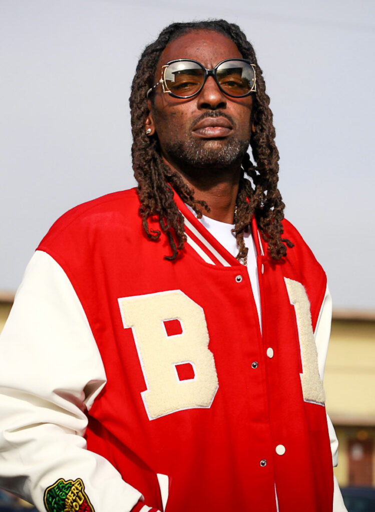 A medium portrait of Cornelius Wills outdoors, looking directly at the camera with a neutral expression. He has long dreadlocks and is wearing large, gold-rimmed sunglasses. He is dressed in a red and white varsity-style jacket with a large cream-colored letter B on the front and a circular patch on the sleeve. The background is a soft-focus view of a building under a clear, bright sky.