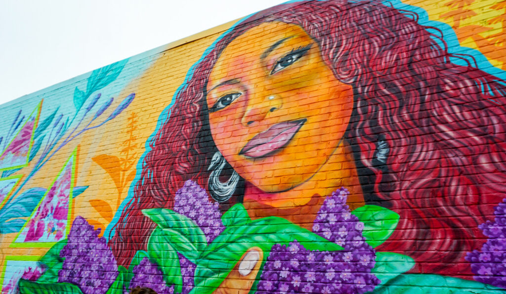 A vibrant outdoor mural painted on a brick wall features a large-scale portrait of a woman with warm, golden skin and long, flowing wavy reddish-brown hair. 