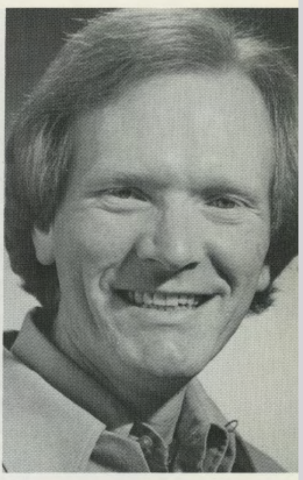 A black and white portrait of Laurence W. Jaquith, Former Professor of Theatre Arts. He is shown from the shoulders up, smiling at the camera with a friendly expression. He has light-colored hair styled in a feathered look and is wearing a button-down shirt with the collar visible. The background is a simple, neutral studio backdrop.