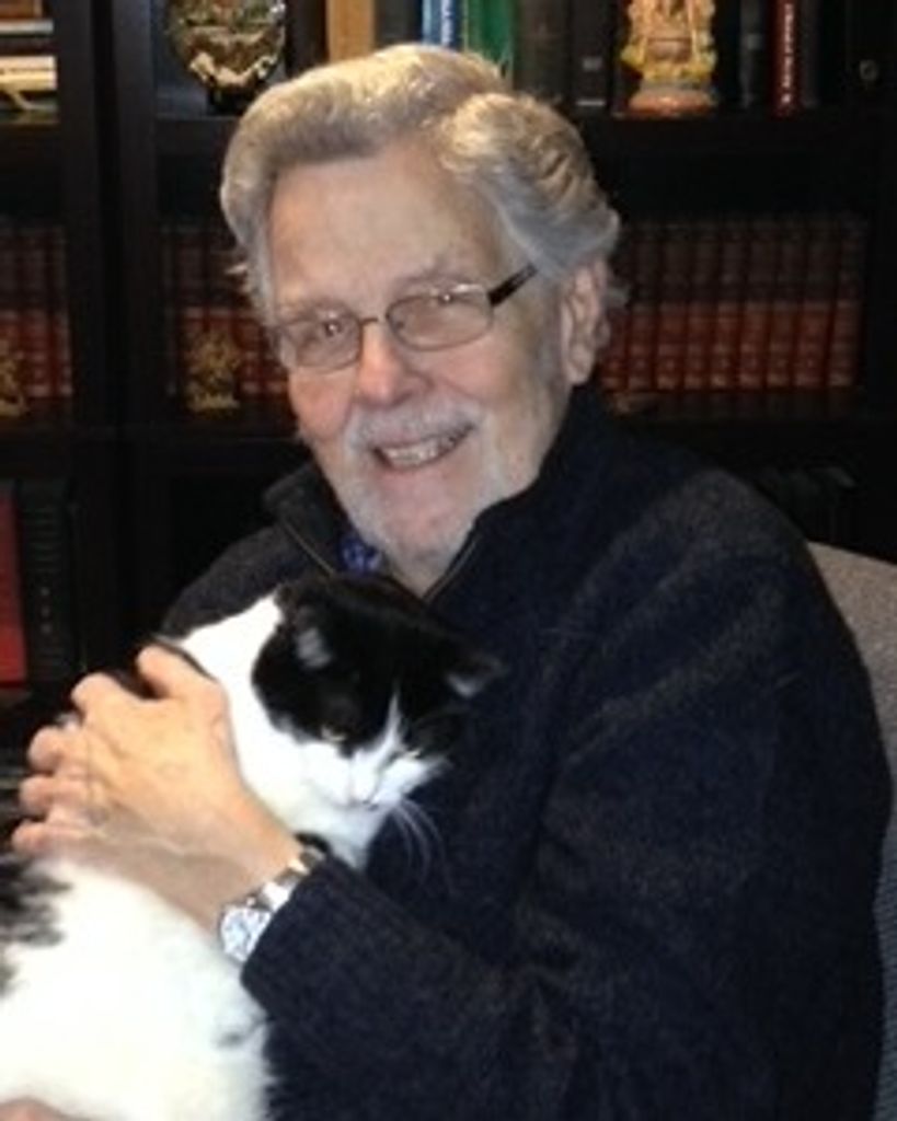 This portrait features John Mark Thompson, an elderly man with thick, wavy grey hair and a gentle smile, wearing rectangular glasses and a dark blue zip-up sweater. He is seated in front of a bookshelf filled with numerous volumes, holding a black and white cat close to his chest. The man is looking directly at the camera, and his left hand, wearing a silver watch, is visible as he cradles the cat.