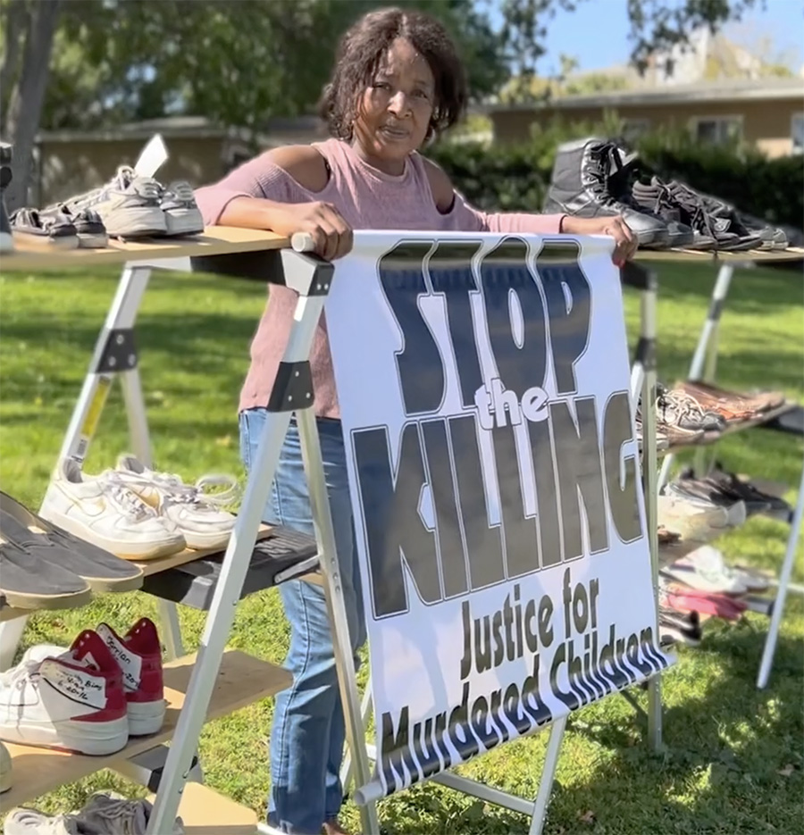 Lawanda Hawkins stands outdoors behind a large white sign that reads STOP the KILLING Justice for Murdered Children in bold black lettering. She is leaning against a temporary shelving unit made of wooden planks and step ladders, which displays several pairs of shoes, some with writing on them. The background shows a grassy lawn and residential buildings under bright daylight.