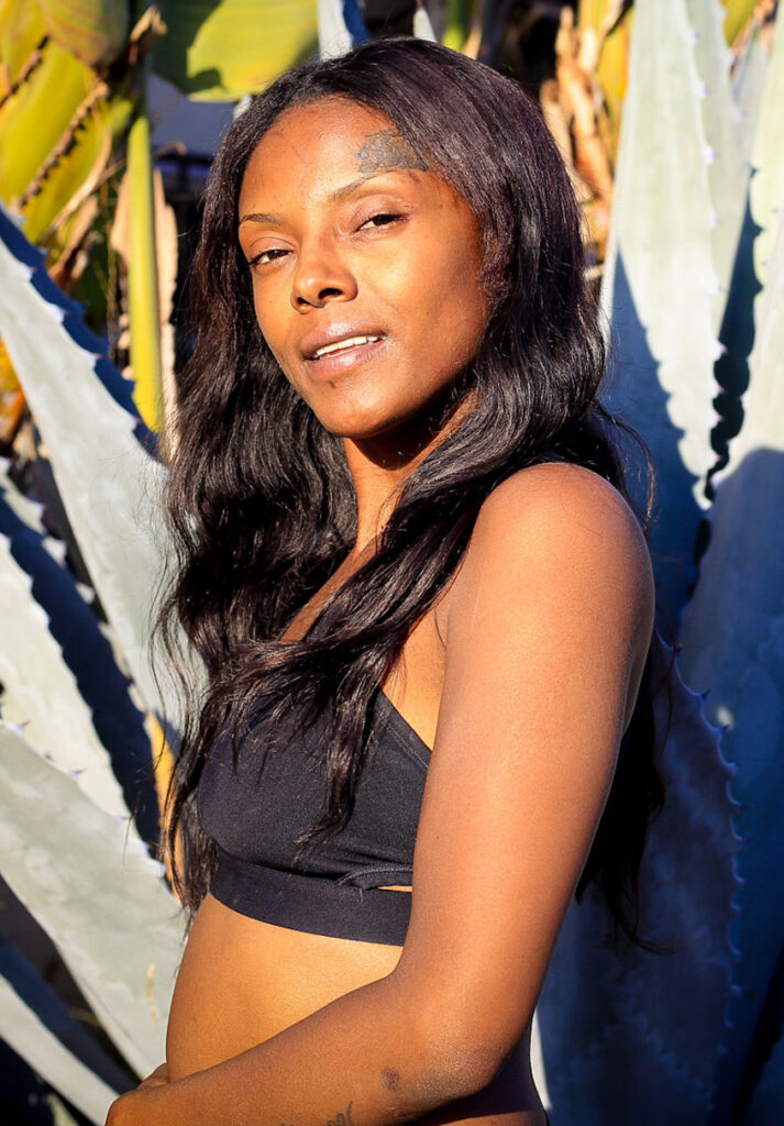 A profile portrait of Neci McKinney, community resident, outdoors in the bright sunlight. She is wearing a black sports bra and has long, wavy dark hair. She looks toward the camera with a gentle expression. In the background are large, blue-green agave plants with sharp spines and some blurred green banana leaves, with strong sunlight casting defined shadows.