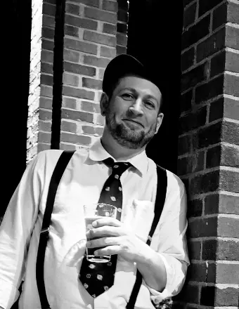 A black and white portrait of Timothy Bayne Northrup, who is leaning against a brick pillar while holding a glass. He is wearing a light-colored button-down shirt, a dark patterned necktie, suspenders, and a dark cap. He has a slight smile and a trimmed beard, looking directly toward the camera.