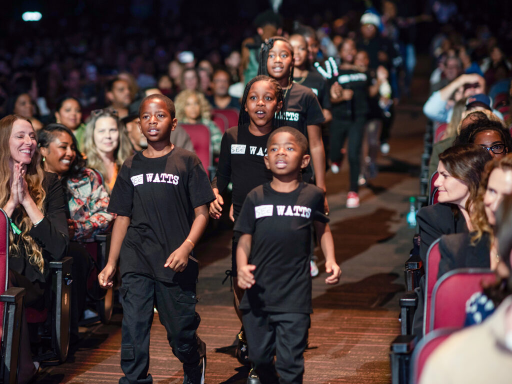 A group of young choir students wearing black t-shirts that say SEE WATTS walk down the aisle of the Peacock Theatre. 