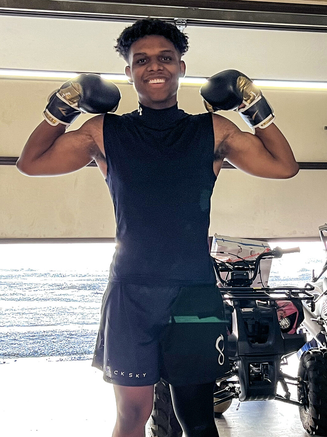 A full-length portrait of a smiling Sean Reynolds standing in a garage. He is wearing black boxing gloves with gold accents, held up near his head in a flexed pose. He is dressed in a black sleeveless athletic shirt, black shorts, and a single black compression legging on his left leg. In the background, an ATV is parked near the open garage door, which looks out onto a bright, sunlit area.