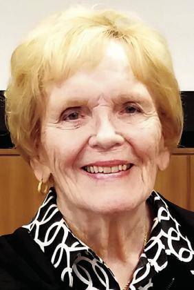 A close-up portrait of Shirley Jo Barron, Former Director for Health Services, a woman with short, blonde hair and a warm smile. She is wearing a black top with a white-and-black patterned collar, a gold necklace, and small gold hoop earrings. The background features a wood-paneled wall and a white screen.