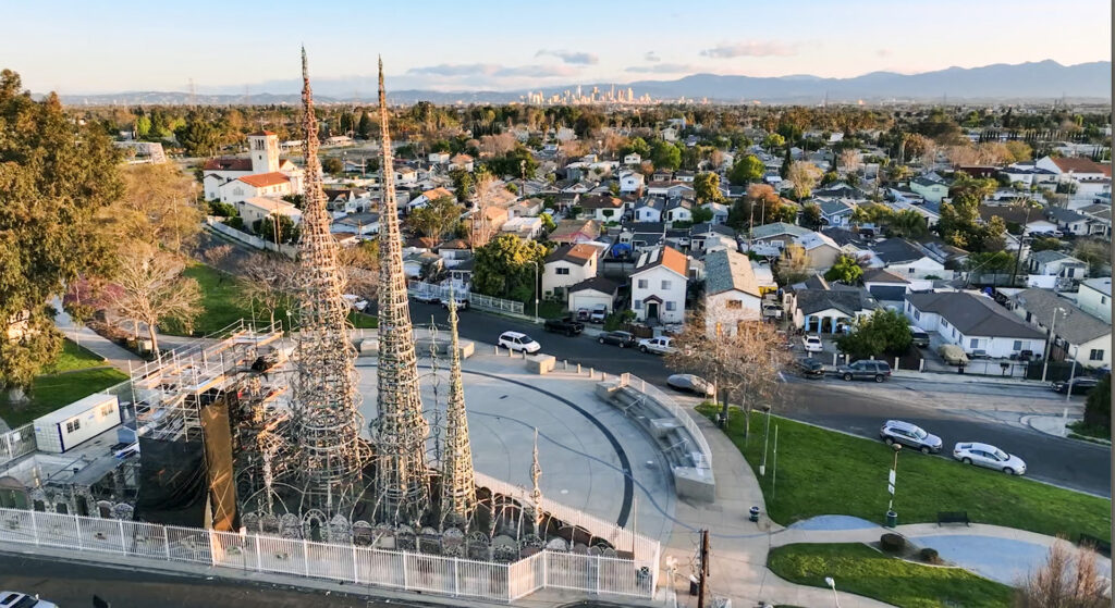 An aerial view of the Watts Towers in Los Angeles, showing the tall, intricate mosaic-covered spires rising above a circular plaza. The surrounding neighborhood consists of residential streets and single-family homes, with a white church visible in the distance. In the far background, the downtown Los Angeles skyline and mountain ranges are visible under a clear, late-afternoon sky.
