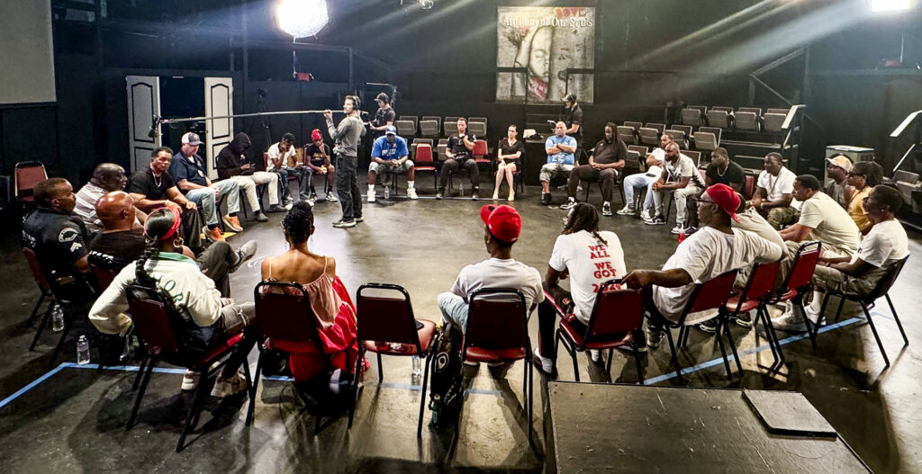 A wide-angle, high-angle shot of a large group of people sitting in a wide circle of red chairs in a darkened studio or community space. A film crew member stands in the center of the circle holding a boom microphone, while another crew member is visible in the background near production equipment. On the back wall, a large poster reads Anthem of Our Souls. The participants are engaged in a group discussion under bright studio lights, creating an atmosphere of serious and focused conversation.